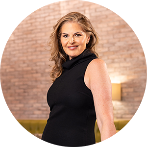 A woman with long, wavy blond hair wearing a sleeveless black dress stands indoors at a Summerlin restaurant in Las Vegas, posing in front of a light-colored brick wall and green bench while smiling at the camera.