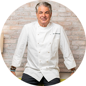 A man with gray hair wearing a white chef’s coat sits in front of a light-colored brick wall, looking at the camera with a relaxed expression, capturing the essence of a renowned Summerlin restaurant in Las Vegas.