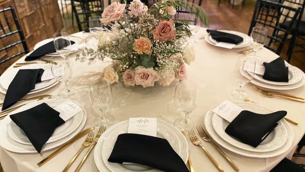 A round table is set for six at this Summerlin restaurant, with white plates, black napkins, gold cutlery, clear glasses, printed menus, and a floral centerpiece featuring pink, peach, and cream flowers with greenery.