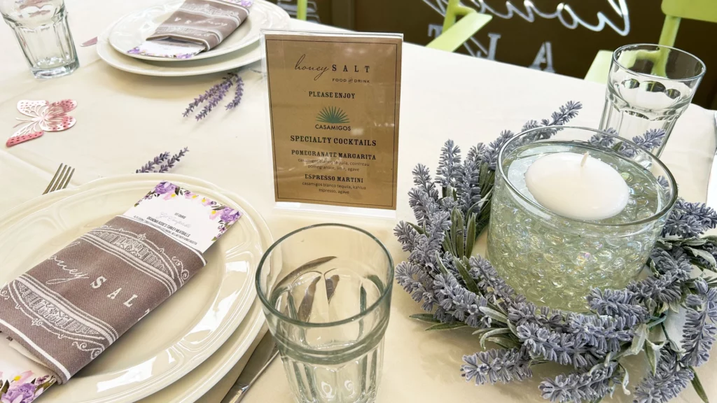 A dining table is set with plates, cutlery, a napkin with honey SALT written on it, a drink menu in a stand, and a glass bowl with a white candle surrounded by artificial lavender flowers in this stylish Summerlin restaurant.