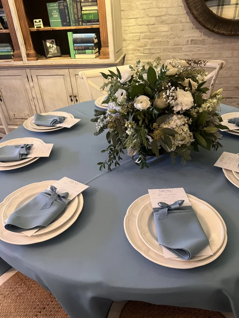 A round dining table at a Summerlin restaurant is covered with a light blue tablecloth and set with six white plates, blue napkins, and printed menus. A large floral arrangement with white flowers and greenery sits in the center.