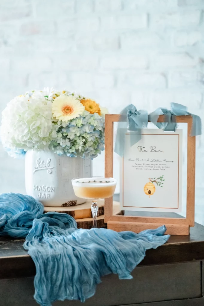 A wooden frame with a bar menu sits next to a white vase of fresh flowers, a cocktail glass, and a blue scarf, all arranged on a table at a stylish Summerlin restaurant against a light brick wall.