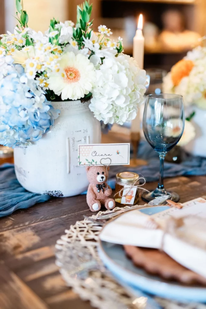 A table setting in a Summerlin restaurant with a blue wine glass, plate, woven placemat, large white vase of flowers, lit candle, small teddy bear, honey jar, and a name card that says Chiara.