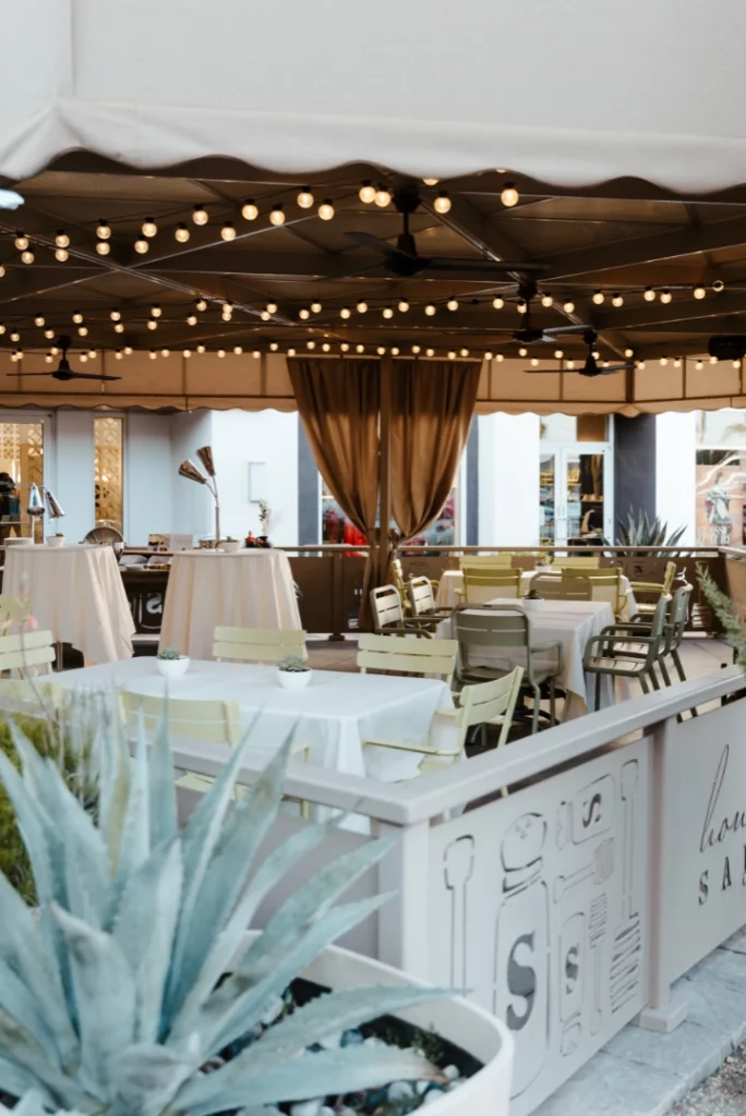 Outdoor restaurant patio in Summerlin with empty tables and chairs under a canopy adorned with string lights. Decorative plants line the foreground, while tan curtains complete this inviting Las Vegas dining area.