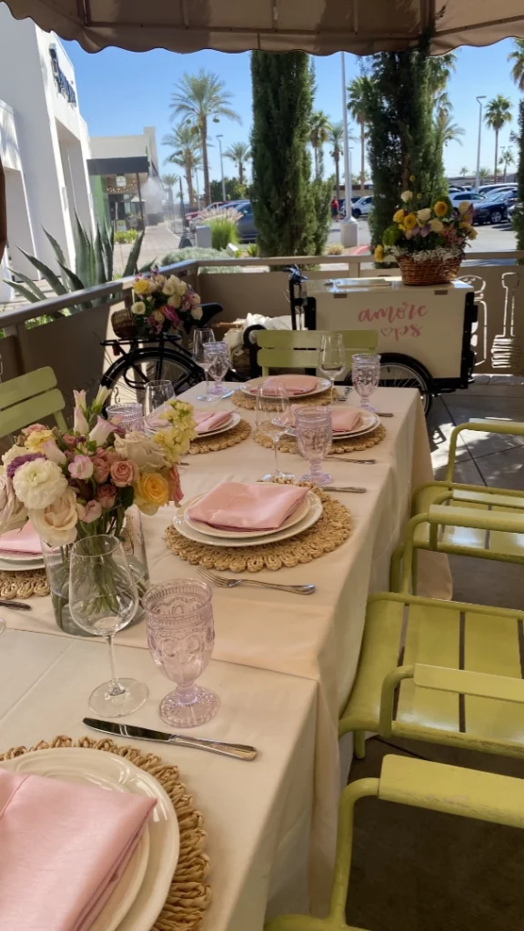A patio table at a restaurant in Summerlin is set for a meal with six place settings, each with a pink napkin, glassware, and floral centerpiece. Green chairs surround the table, and potted flowers decorate the area near Las Vegas palm trees.