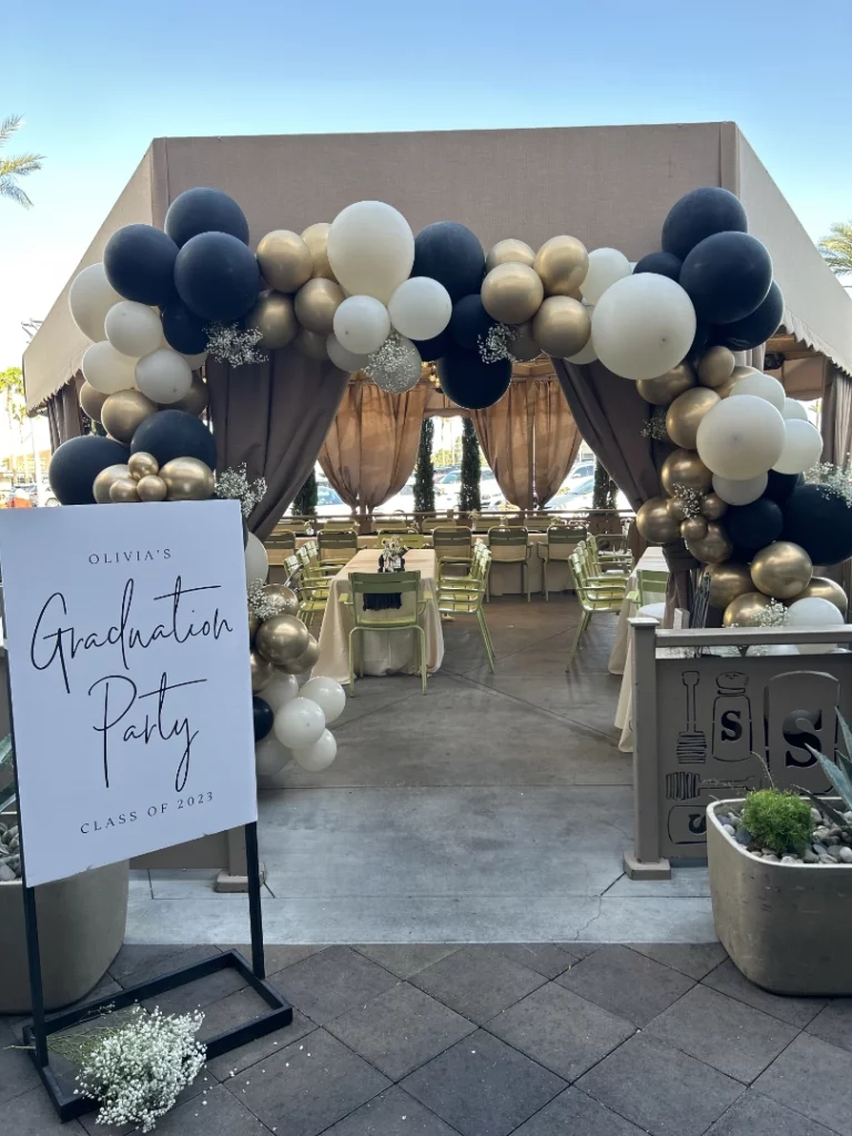 A decorated outdoor tent at a Summerlin restaurant features gold, black, and white balloons forming an arch over the entrance. A sign reads Olivia's Graduation Party, Class of 2023. Tables and chairs are set up inside the tent.