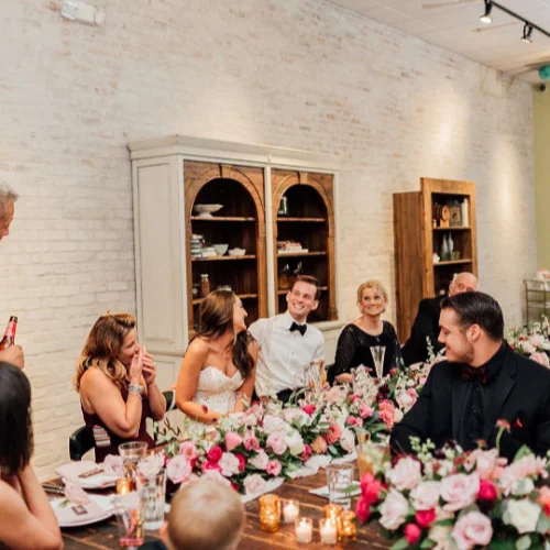 A wedding group sit at a long table decorated with pink and white flowers and candles in a warmly lit Honey Salt restaurant.