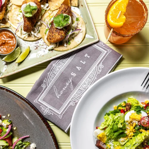 Overhead view of a table at Honey Salt, A Summerlin restaurant with fish tacos, a salad, a drink with an orange slice.